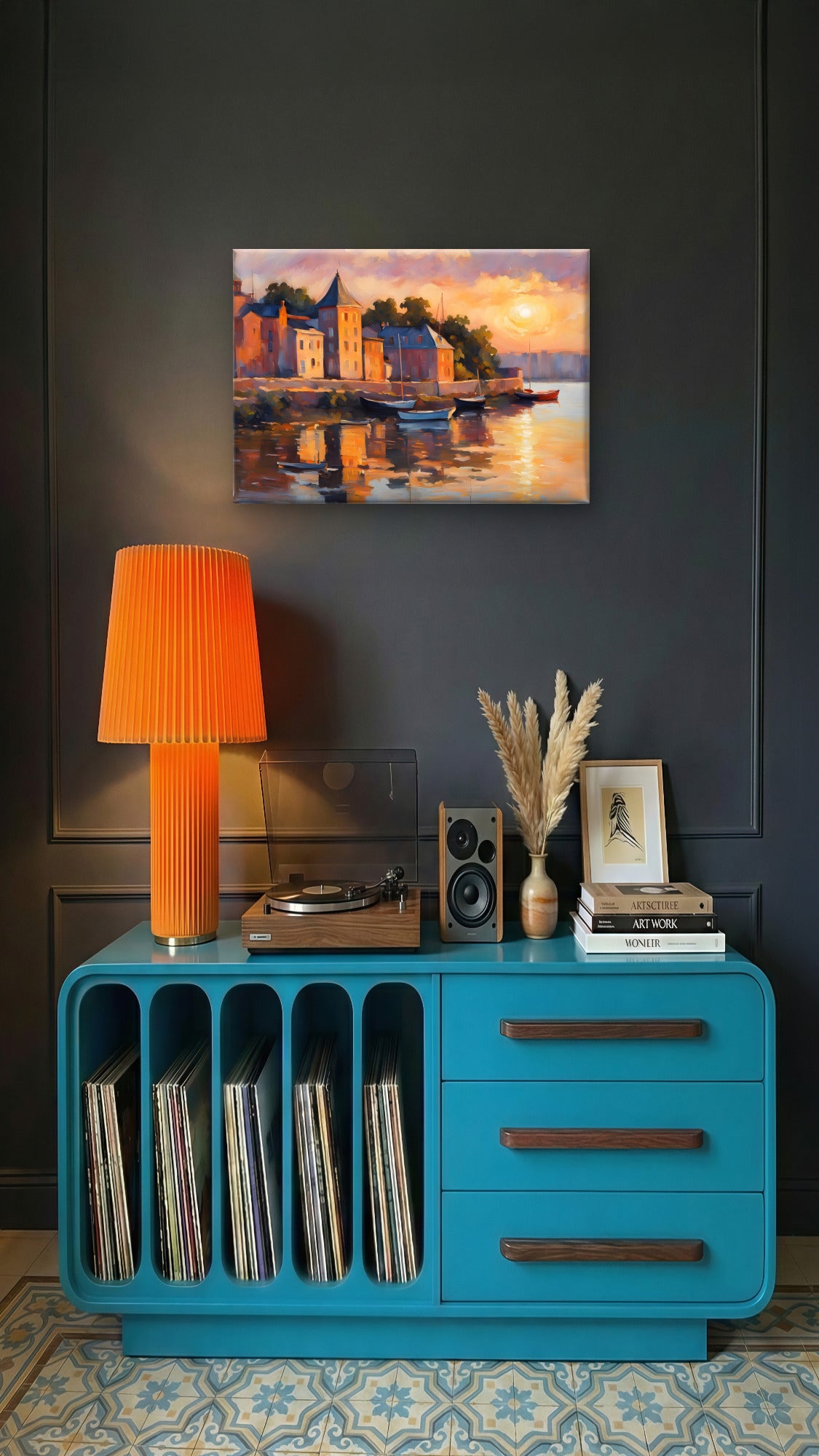 A bright turquoise sideboard cabinet styled in a modern living room setting, featuring a pleated orange lamp, a vinyl record player, speakers, books, and dried grass in a vase. Above the cabinet, a colorful painting of a coastal village at sunset hangs on a dark gray paneled wall.