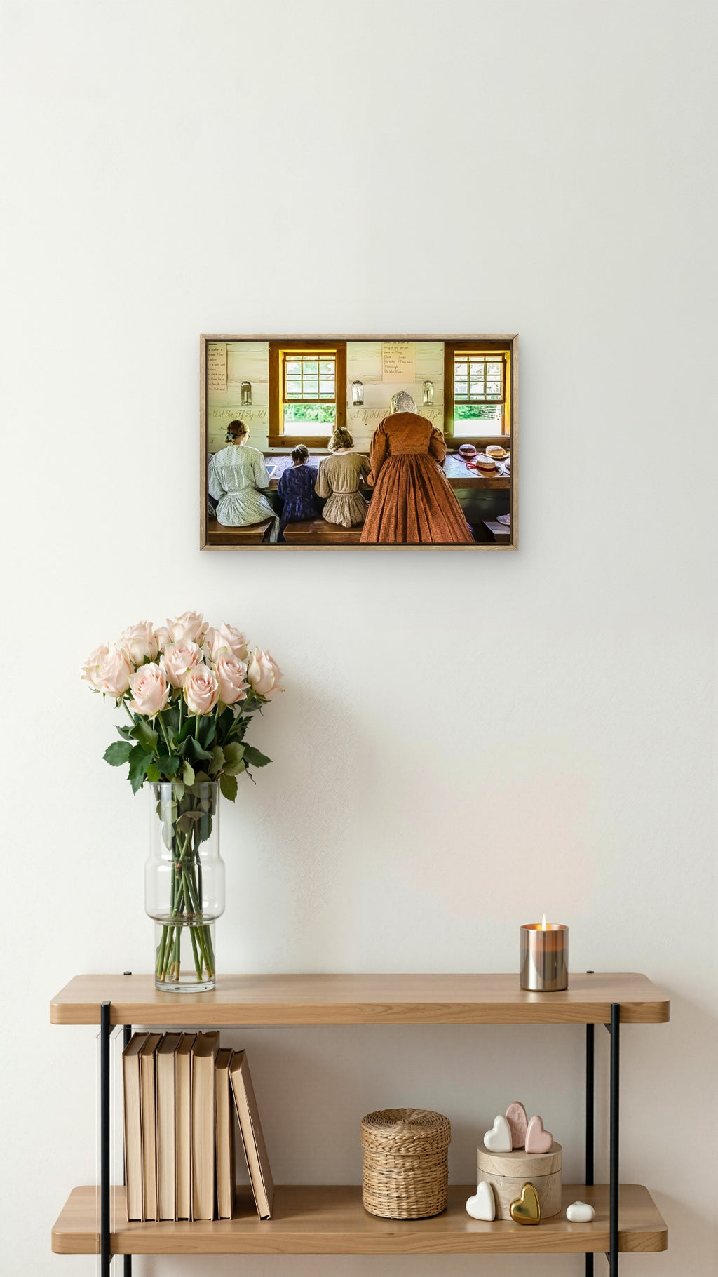 A framed art print depicting a vintage interior scene with a woman in a brown dress and others seated by windows, displayed on a white wall above a wooden shelf with books, a vase of pink flowers, and a candle.