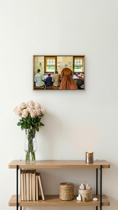 A framed art print depicting a vintage interior scene with a woman in a brown dress and others seated by windows, displayed on a white wall above a wooden shelf with books, a vase of pink flowers, and a candle.
