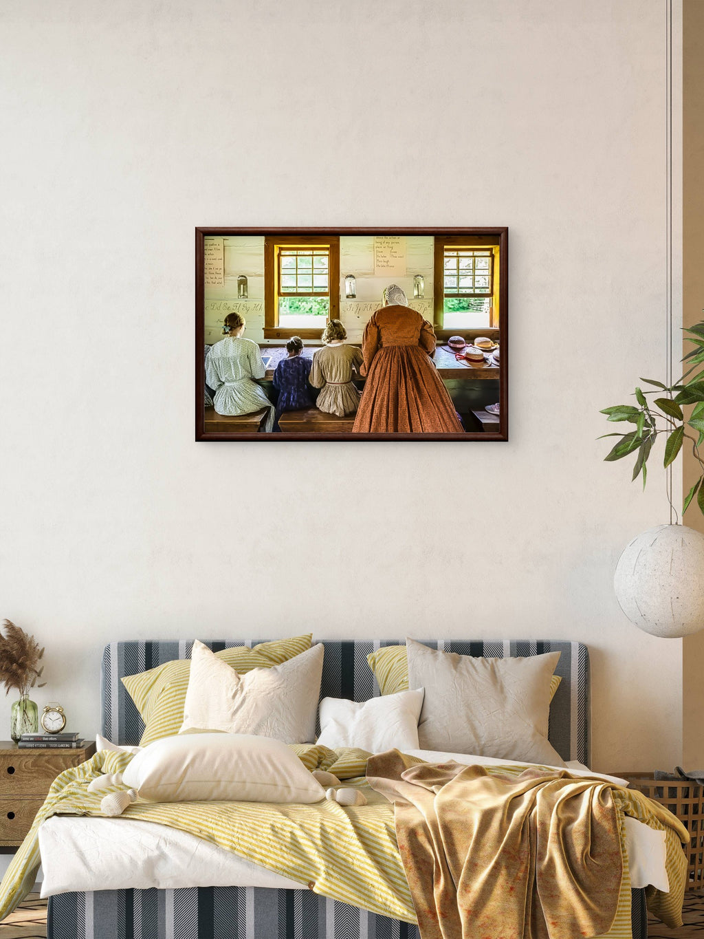 A framed art print depicting a vintage interior scene with women at a table, displayed on a beige wall above a bed with yellow and white bedding in a styled bedroom setting.
