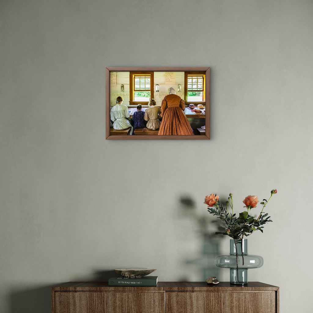 A framed art print depicting a vintage scene of people sewing at windows, displayed on a textured gray wall above a wooden cabinet with a glass vase of flowers.