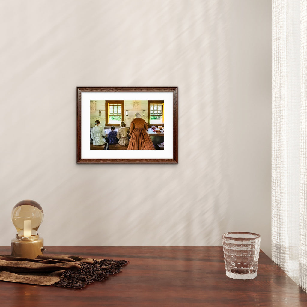 A framed art print depicting a historical scene with figures in a room, styled on a wooden table with a glass, scarf, and lamp against a wall with soft window shadow patterns.