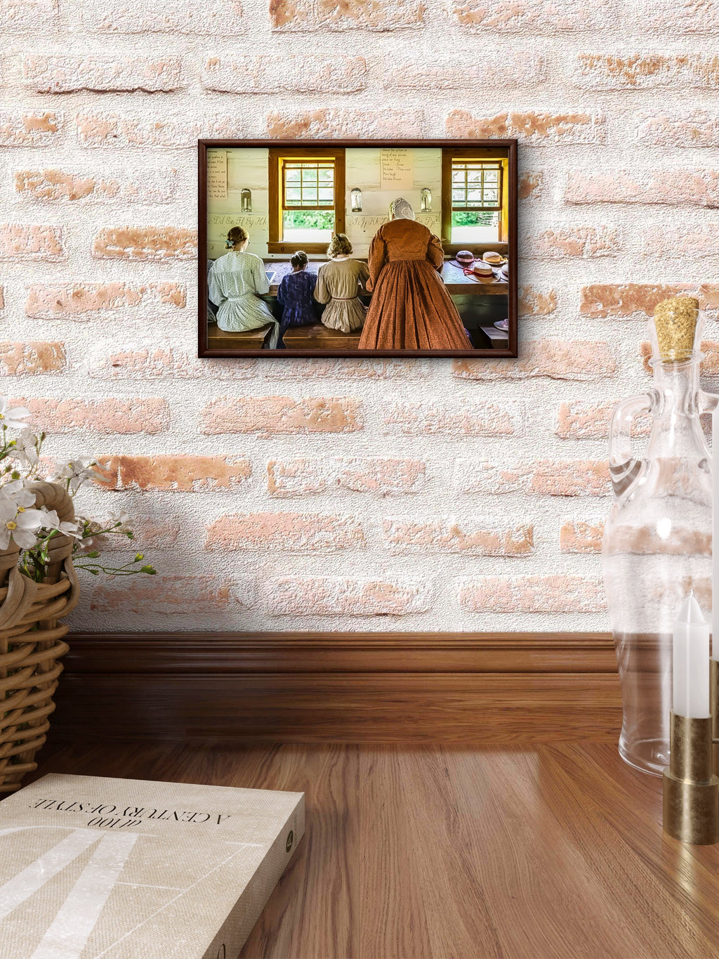 A framed art print depicting a historical scene with women in a kitchen, displayed on a white brick wall above a wooden table with a book and a glass bottle.
