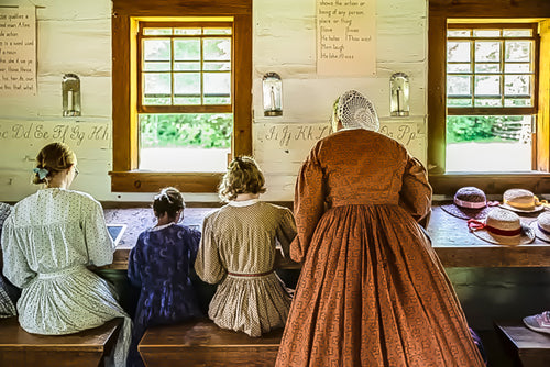 Four people in historical-style period dresses seated and standing in a rustic wooden room with windows, looking toward a counter with pastries and lanterns.