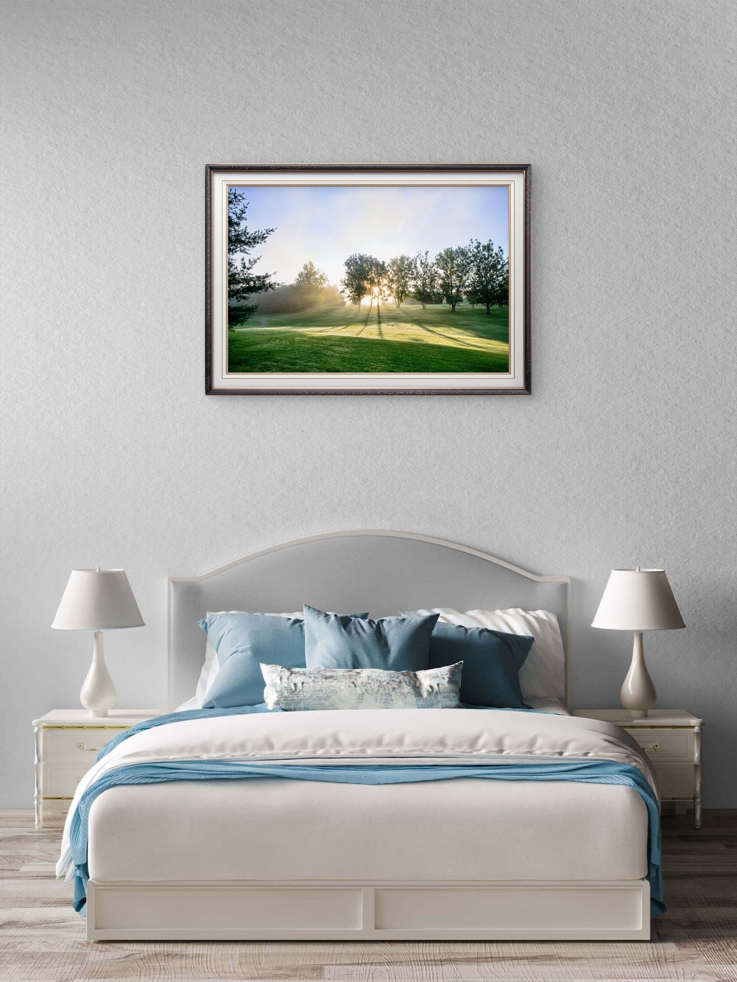 A framed landscape art print of a sunlit golf course is displayed on a textured light gray wall above a neatly made bed with blue and white bedding in a modern bedroom setting.