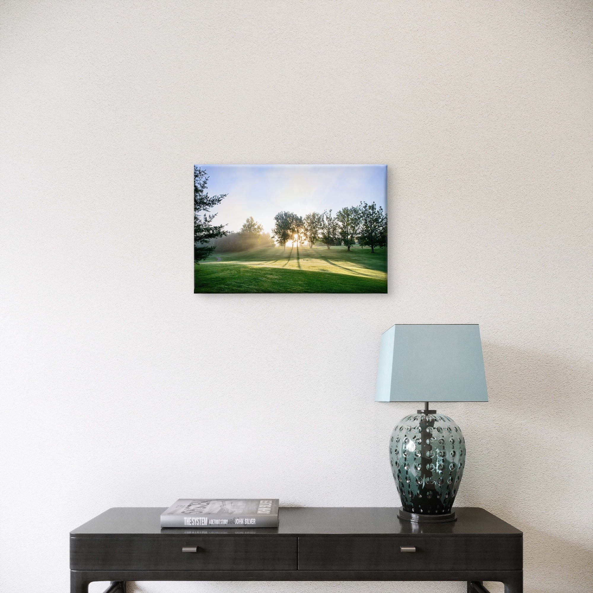 A rectangular wall art print showing a sunlit green field with trees, displayed above a dark console table with a lamp and a book in a real interior setting.