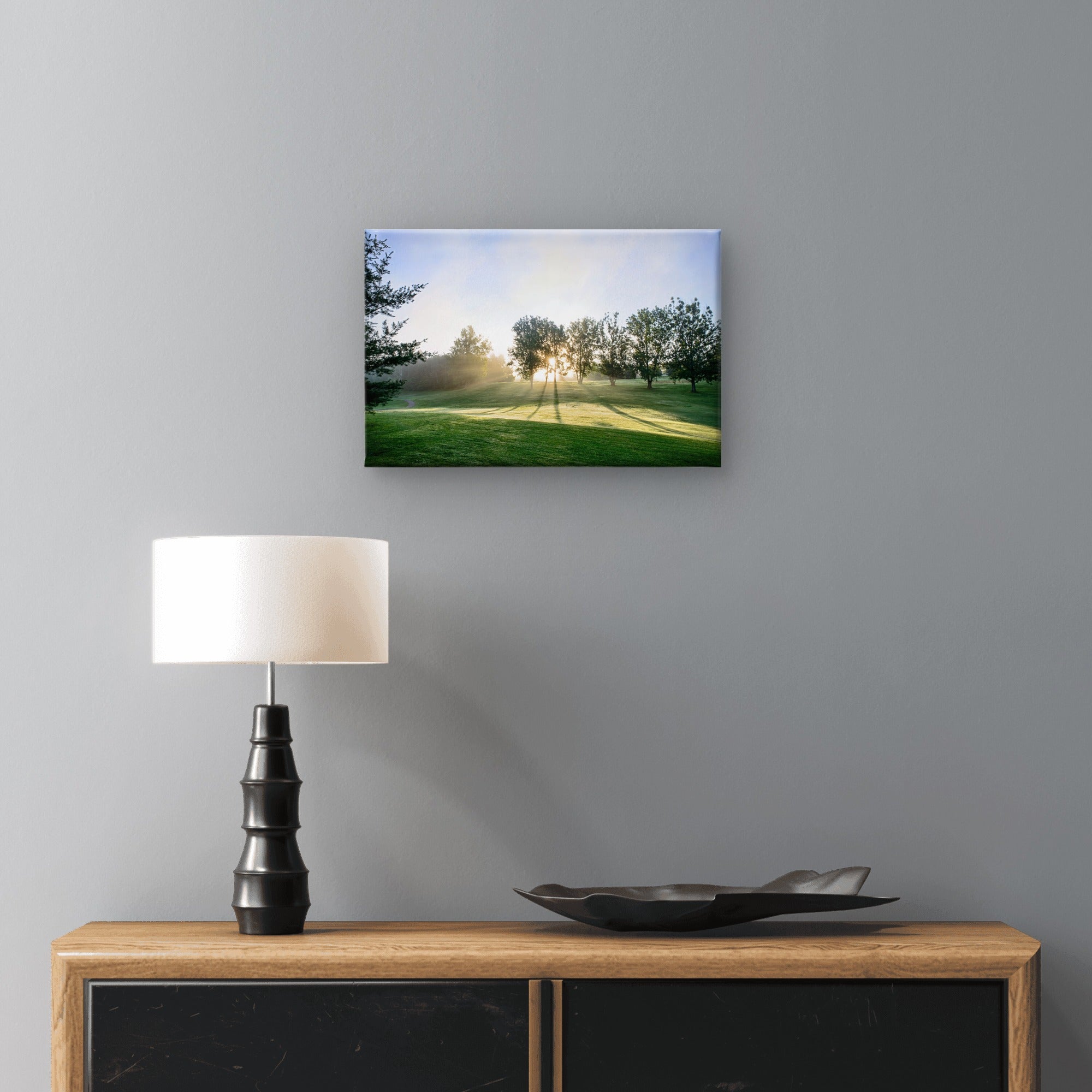A framed wall art print showing a sunlit green landscape with trees, displayed above a wooden console table with a black lamp and a dark tray against a gray wall.