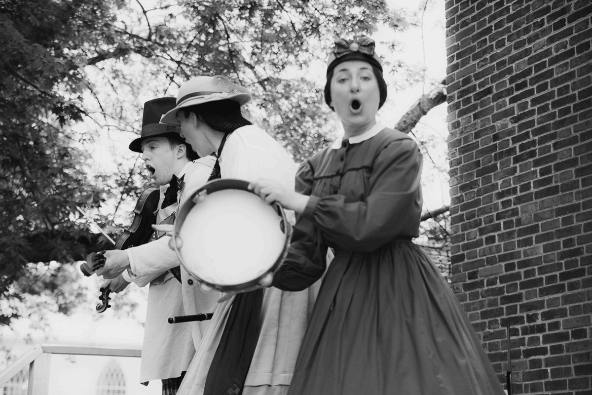 Black and white photograph of three people in historical costume dancing and playing instruments outdoors beside a brick wall.