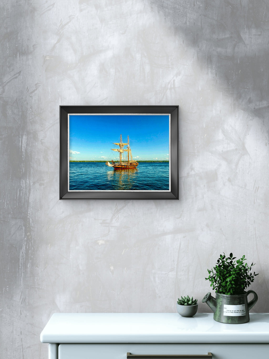 A framed square art print depicting a tall ship on blue water under a clear blue sky, mounted on a textured light gray wall above a white console table with small potted plants.