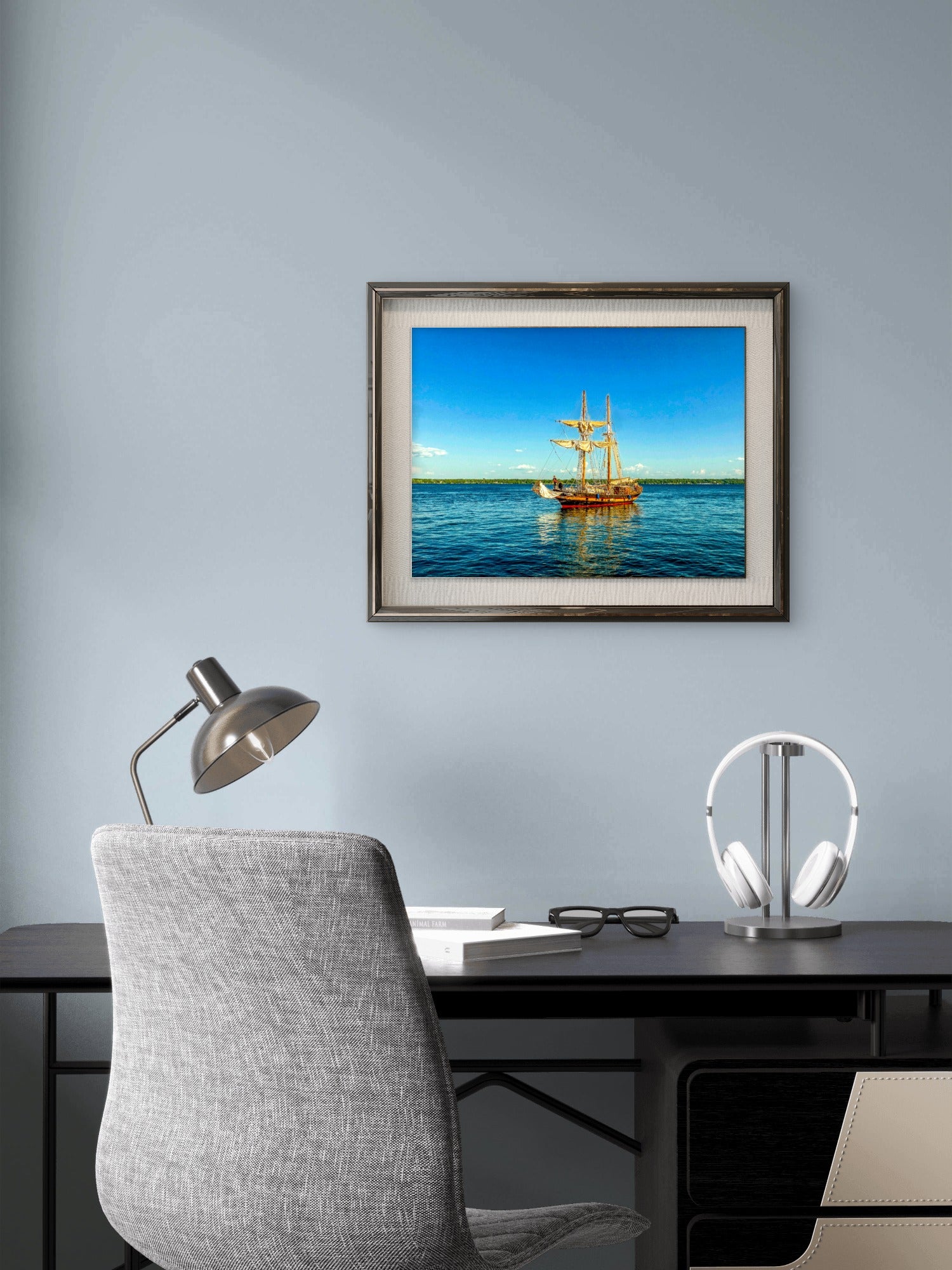 A framed art print of a tall ship sailing on blue water under a clear sky, displayed on a light blue-gray wall above a modern desk setup with a gray chair, silver desk lamp, and white headphones.