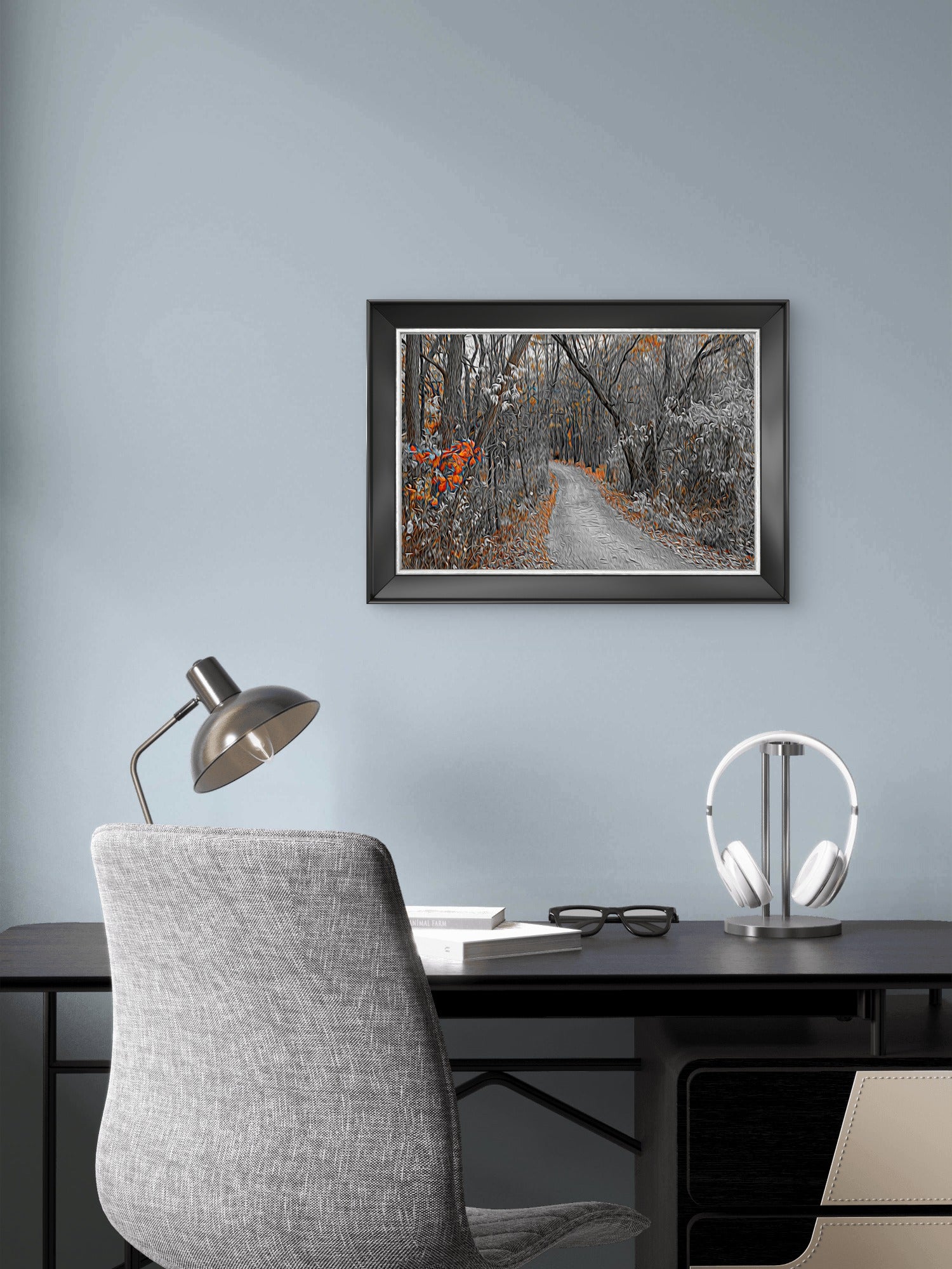 A framed landscape art print of a snowy forest path with orange autumn leaves, displayed on a light blue-gray wall above a modern desk setup with a gray chair, silver desk lamp, and white headphones.